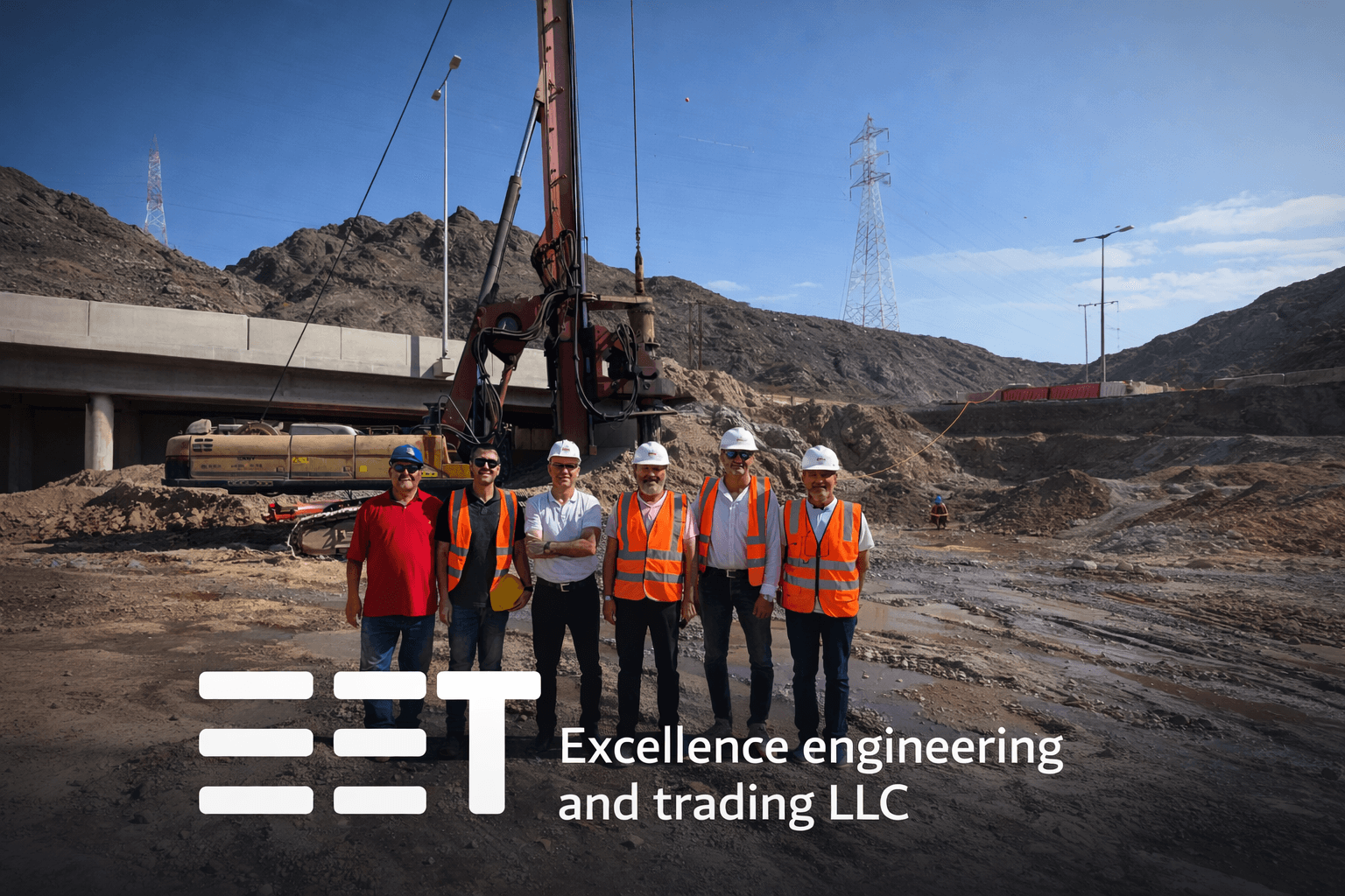 Six engineers in safety gear pose before a drilling rig at a mountainous construction site.
