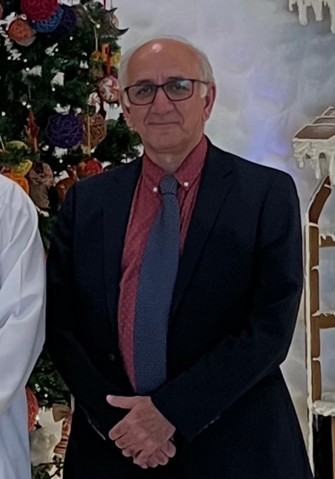 Smiling older man with glasses, dark blazer, and blue tie beside a decorated Christmas tree.