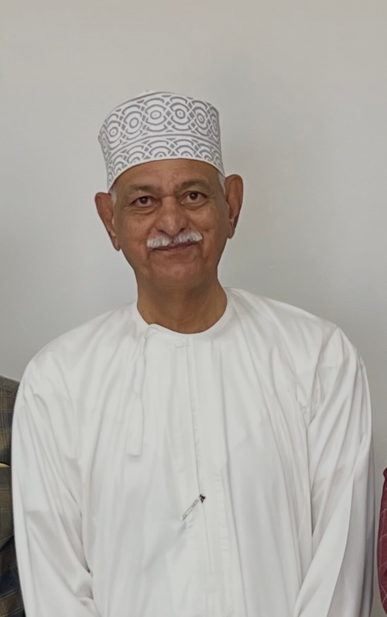 Smiling older man with a white mustache wearing a patterned Omani kumma and white dishdasha.