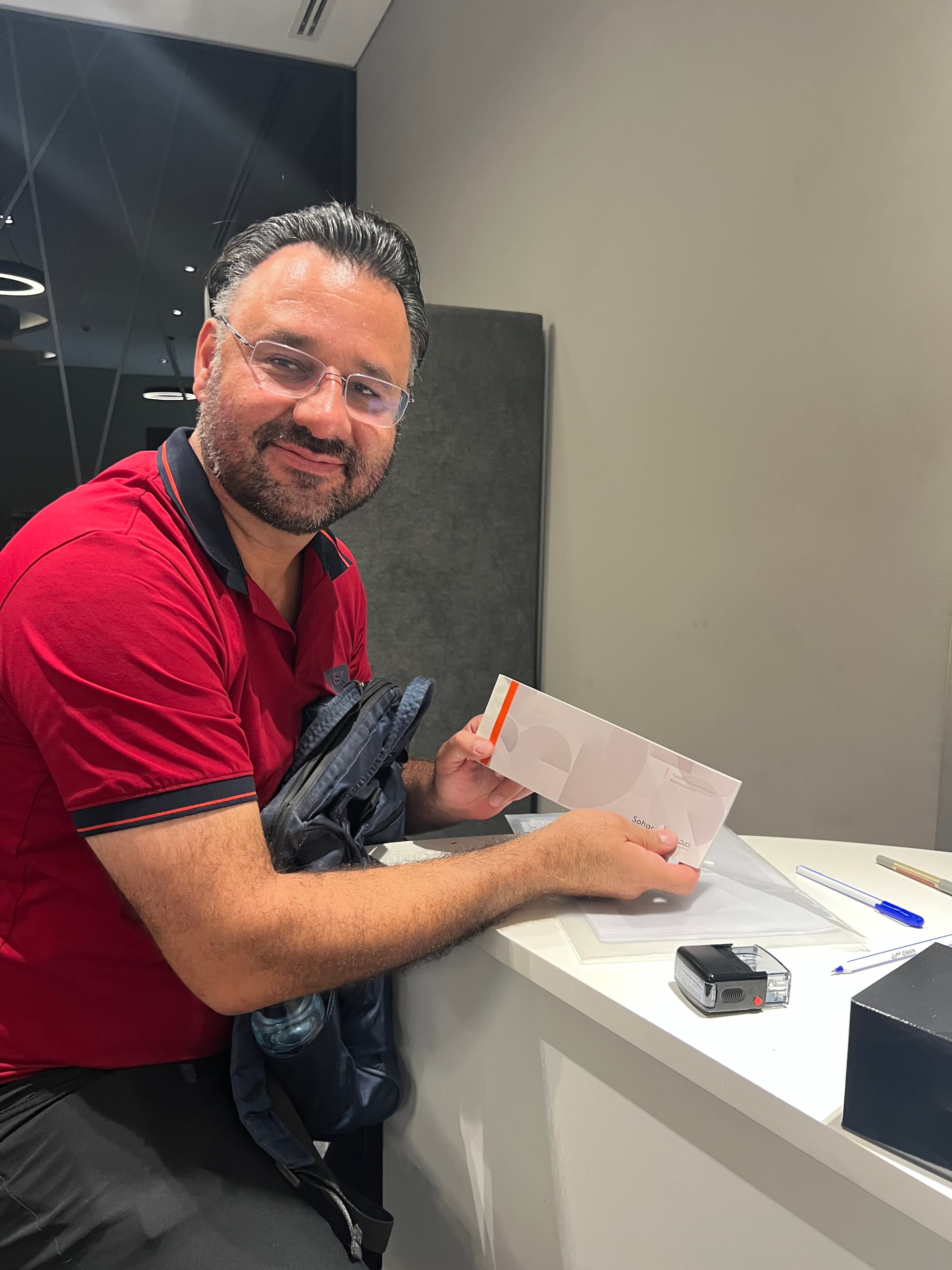 Smiling man in a red polo shirt holds a white Sohar envelope at a desk.