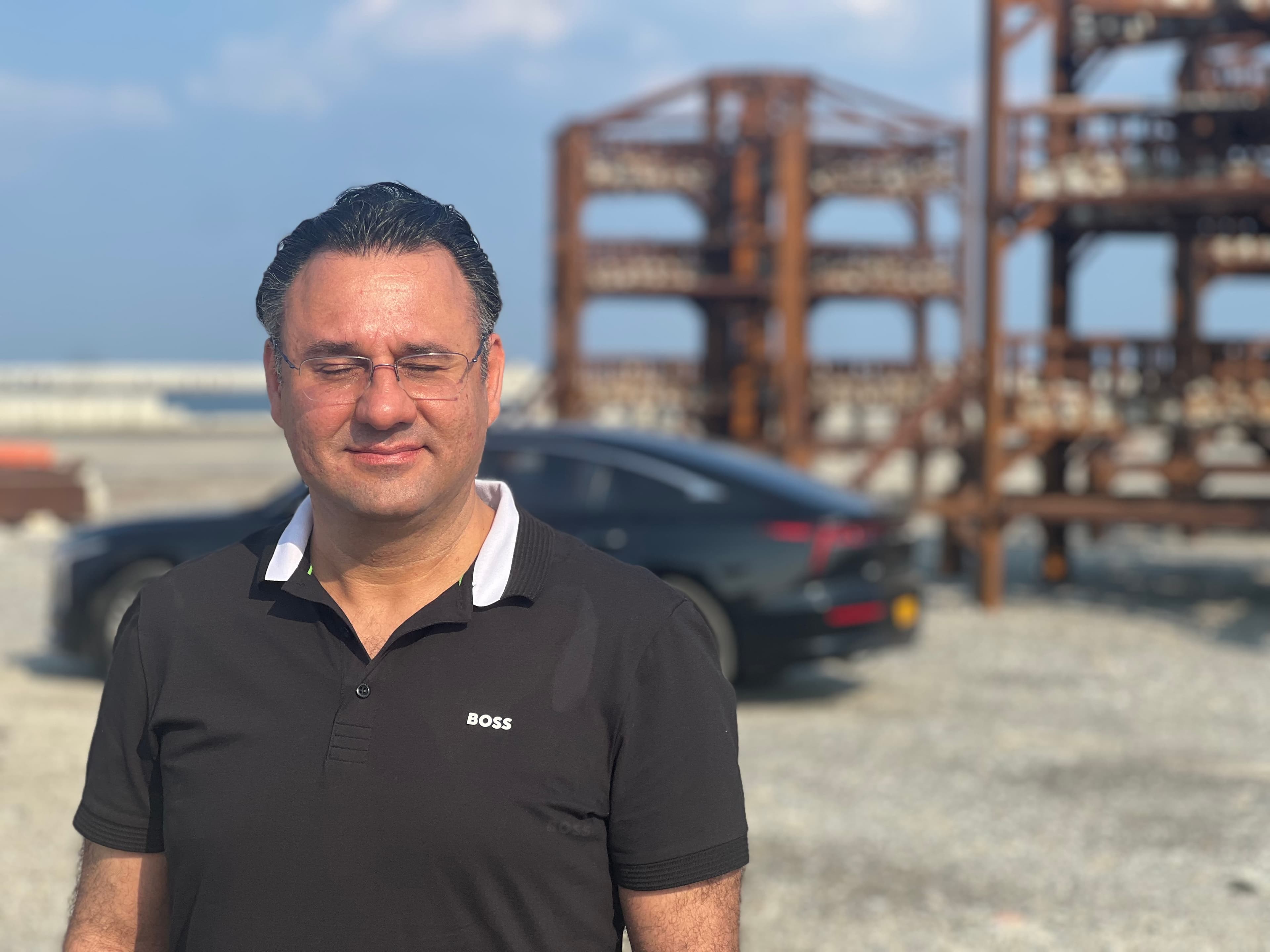 Man with glasses and eyes closed wearing a black BOSS polo shirt near a car.